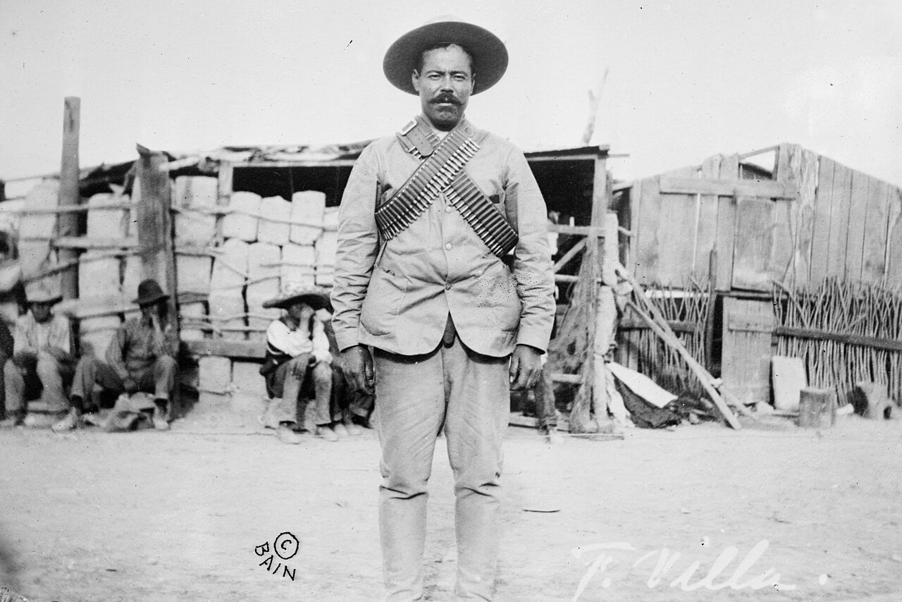 Pancho Villa wearing crossed bandoliers, standing in front of a revolutionary camp
