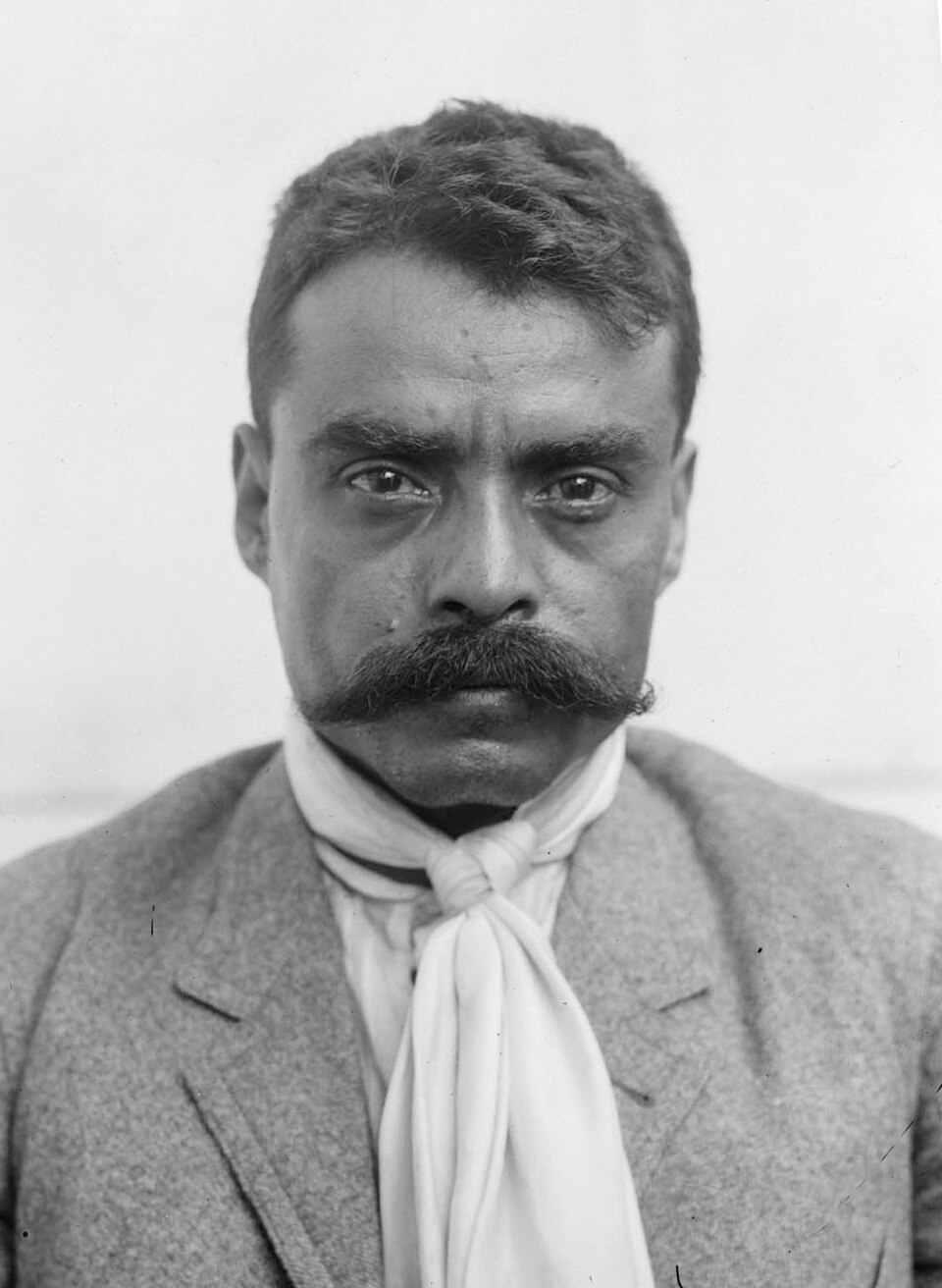 Emiliano Zapata standing in charro clothing with a rifle, sash and sword